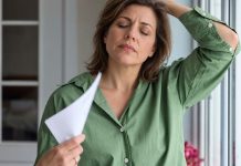Perimenopauzė, simptomai ir kada ji prasideda Perimenopauzė, moteris žaliais marškiniais vėdinasi popieriaus lapu prie lango