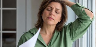 Perimenopauzė, simptomai ir kada ji prasideda Perimenopauzė, moteris žaliais marškiniais vėdinasi popieriaus lapu prie lango