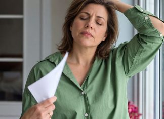 Perimenopauzė, simptomai ir kada ji prasideda Perimenopauzė, moteris žaliais marškiniais vėdinasi popieriaus lapu prie lango