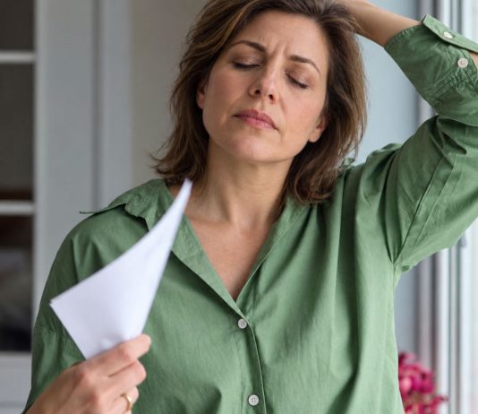 Perimenopauzė, simptomai ir kada ji prasideda Perimenopauzė, moteris žaliais marškiniais vėdinasi popieriaus lapu prie lango