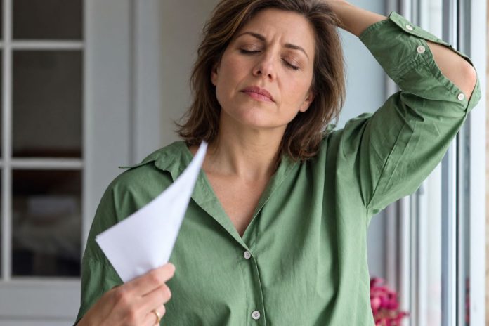 Perimenopauzė, moteris žaliais marškiniais vėdinasi popieriaus lapu prie lango